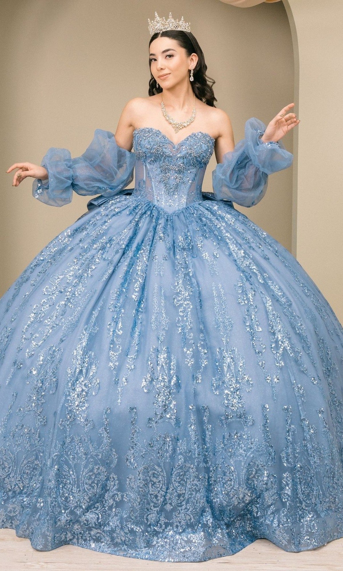 A woman wearing the Quinceanera Dress 8565JQ By Cinderella Couture in Dusty Blue stands indoors, posing with arms raised. The sparkling strapless ball gown features an embellished corset bodice, sheer removable puff sleeves, a crown, and elegant jewelry to complete her look.